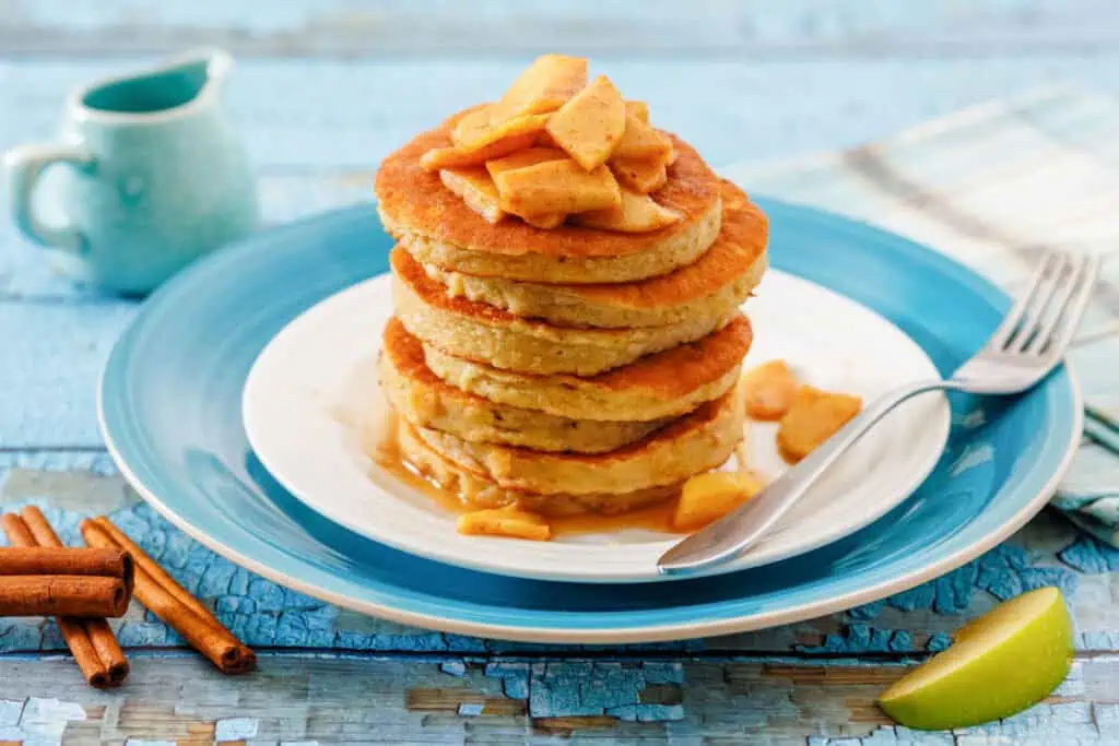 A stack of pancakes topped with cooked apple slices and syrup, served on a white and blue plate with a fork, next to cinnamon sticks and a wedge of green apple.