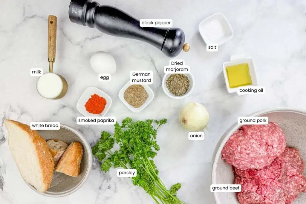 Ingredients for a Frikadellen recipe are laid out on a white surface, including ground pork, ground beef, spices, herbs, bread, milk, egg, onion, oil, salt, and pepper.