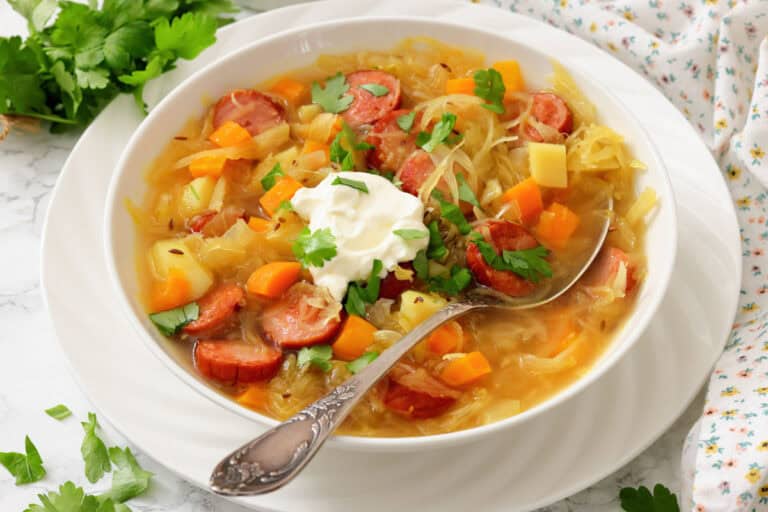 A bowl of cabbage soup with sausage, carrots, potatoes, and fresh herbs, topped with a dollop of sour cream, with a spoon resting in the bowl.