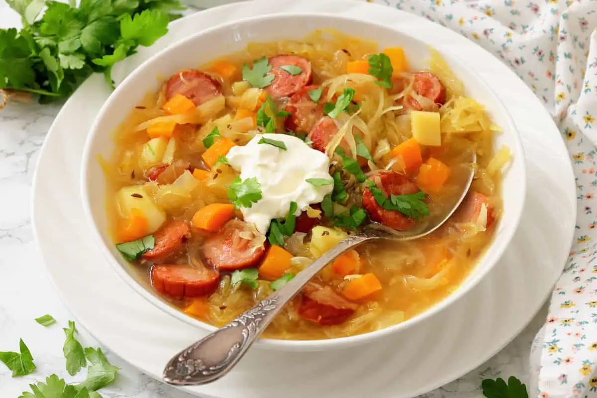 A bowl of soup with sliced sausage, potatoes, carrots, cabbage, garnished with fresh herbs and a dollop of sour cream, with a spoon resting in the bowl.