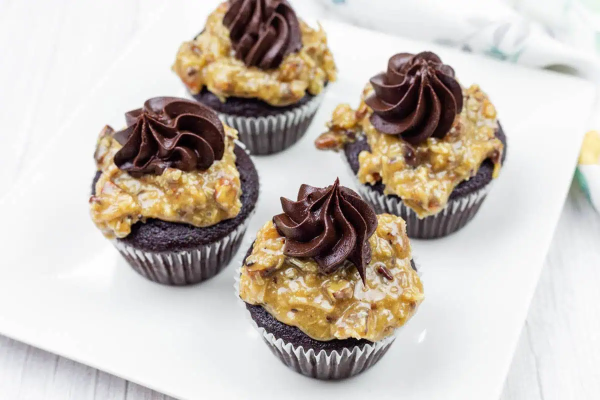 Four German Dark chocolate cupcakes topped with German chocolate coconut-pecan frosting and swirls of chocolate icing, arranged on a white square plate.