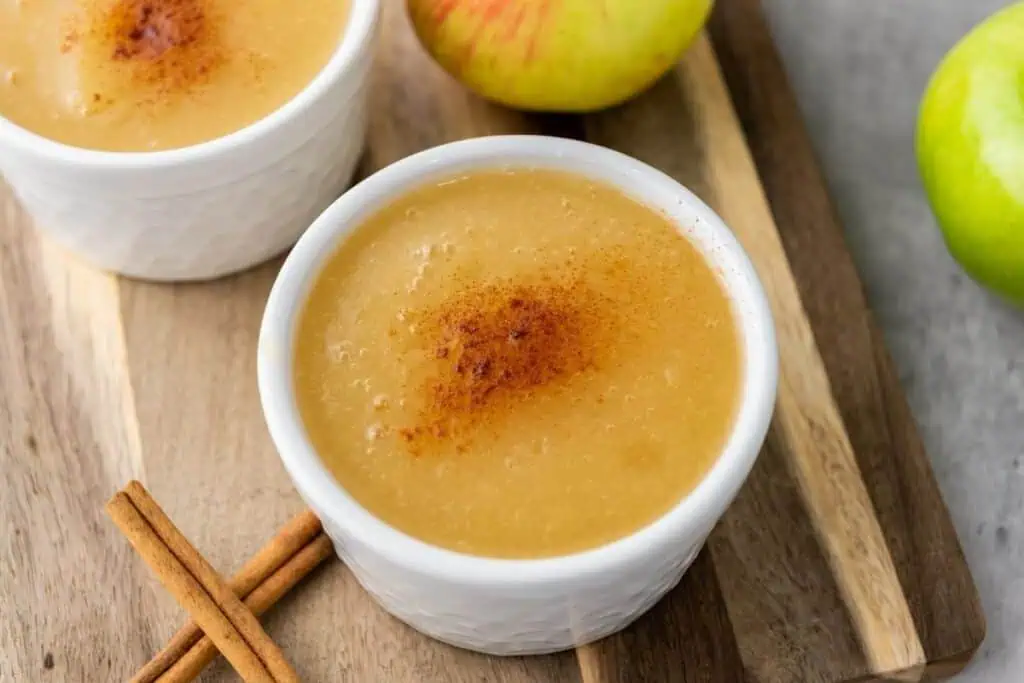 Two white bowls of applesauce topped with ground cinnamon, placed on a wooden board with fresh green apples and cinnamon sticks nearby.