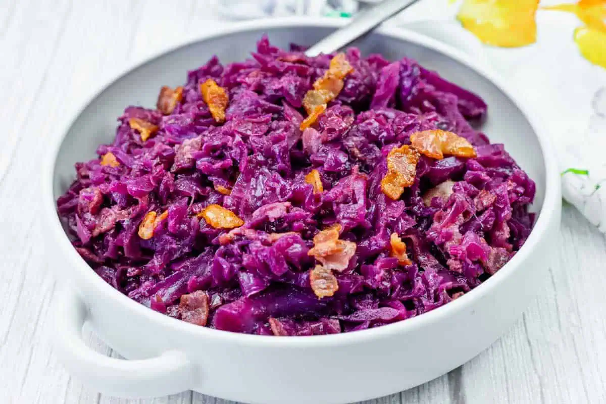 A white bowl filled with cooked red cabbage and pieces of bacon, placed on a light wooden surface.