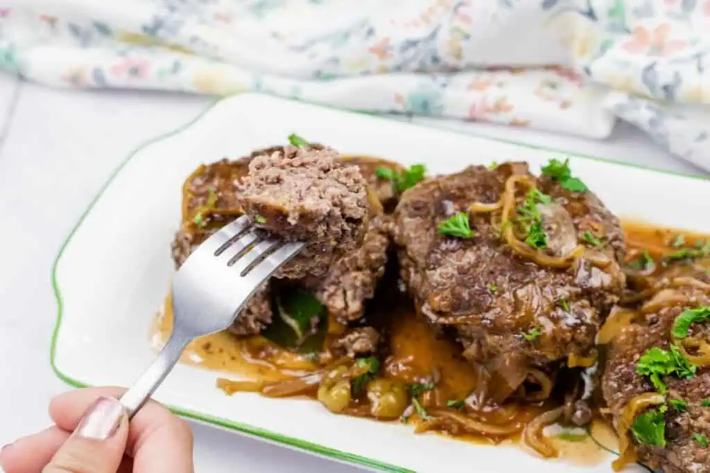 A hand holds a fork with a bite of Salisbury steak over a plate topped with gravy, onions, and parsley garnish.