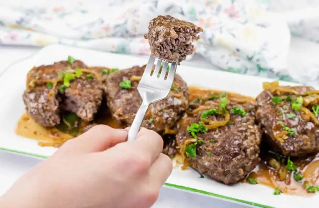 A hand holds a fork with a bite of Salisbury steak above a plate of several steaks topped with gravy and garnished with herbs and onions.