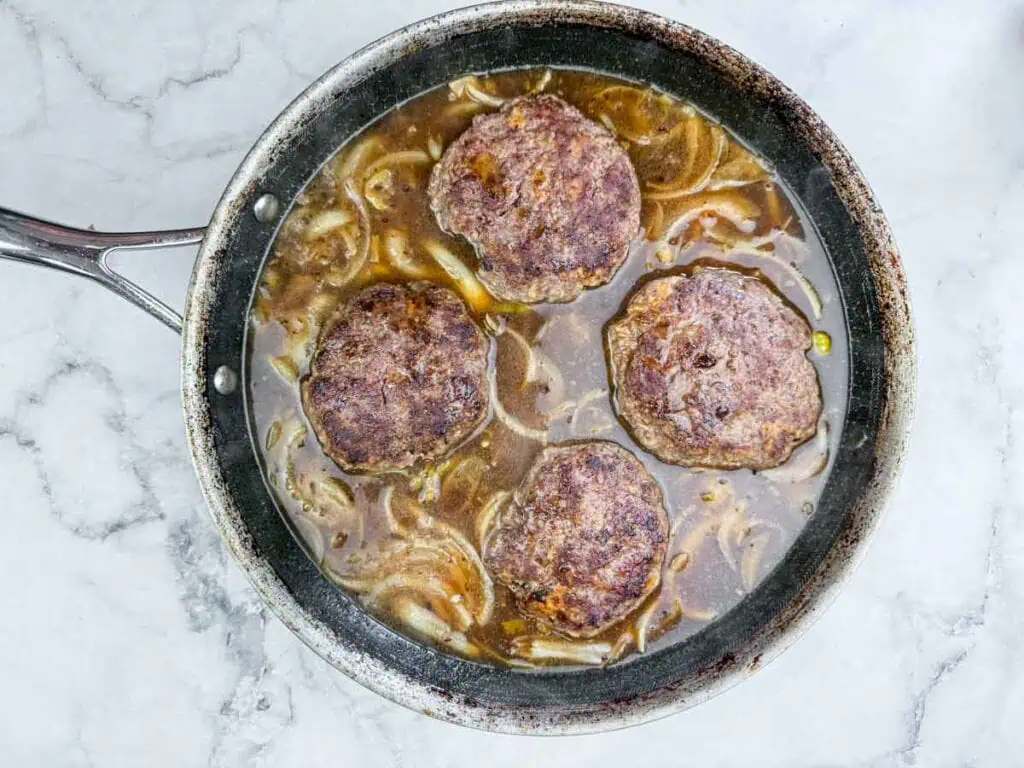 Four cooked hamburger patties simmer in brown onion gravy inside a skillet on a marble countertop.