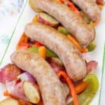 Four cooked sausages are served on a platter with roasted potatoes, red onions, carrots, and red and green bell peppers.
