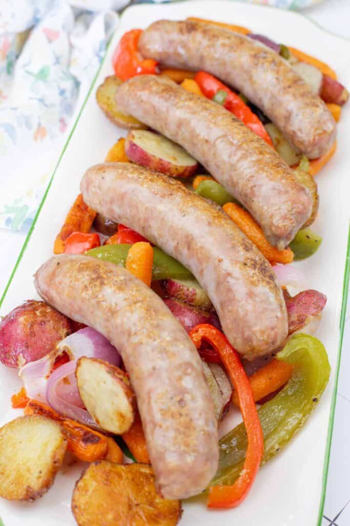 Four cooked sausages are served on a platter with roasted potatoes, red onions, carrots, and red and green bell peppers.