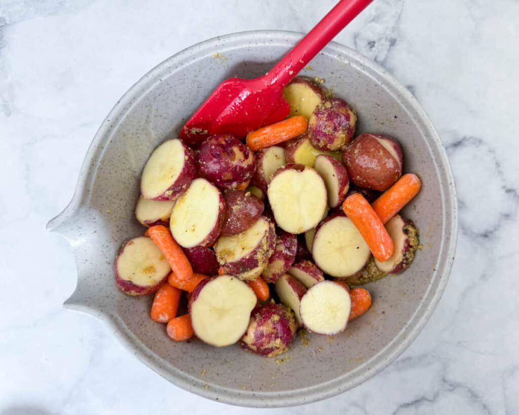 A bowl containing sliced red potatoes and baby carrots being mixed with seasoning using a red spatula on a marble surface.