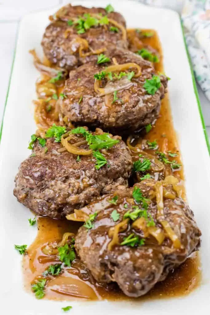 Four cooked hamburger steaks topped with sautéed onions and brown gravy, garnished with chopped parsley, served on a rectangular white platter.