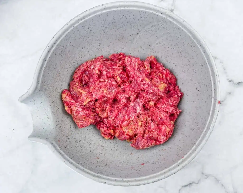 A speckled gray mixing bowl containing raw ground meat mixture on a white marble surface.