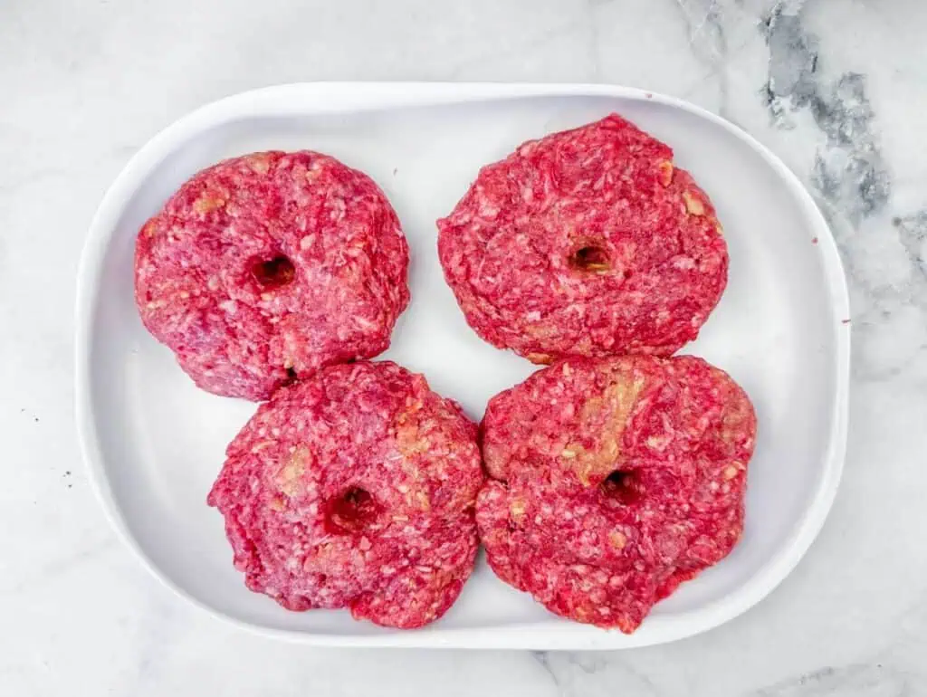 Four raw beef burger patties with holes in the center are arranged on a white tray atop a marble surface.