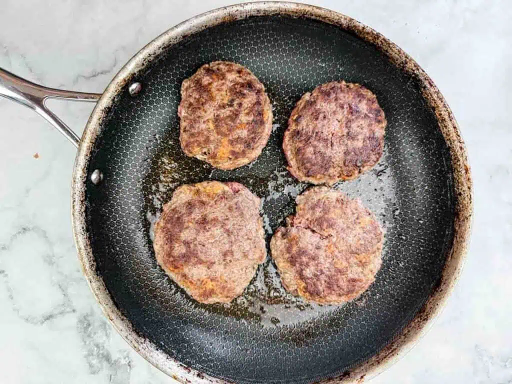 Four browned burger patties cooking in a frying pan on a stovetop.