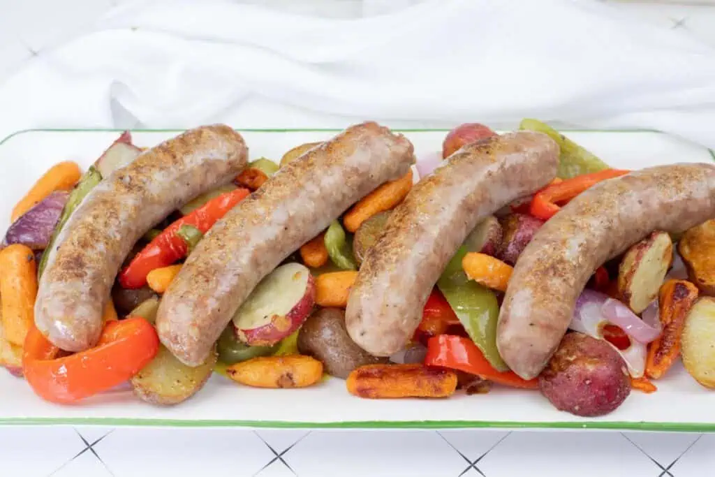 Four cooked sausages are served on a platter with roasted potatoes, carrots, red peppers, green peppers, and onions.