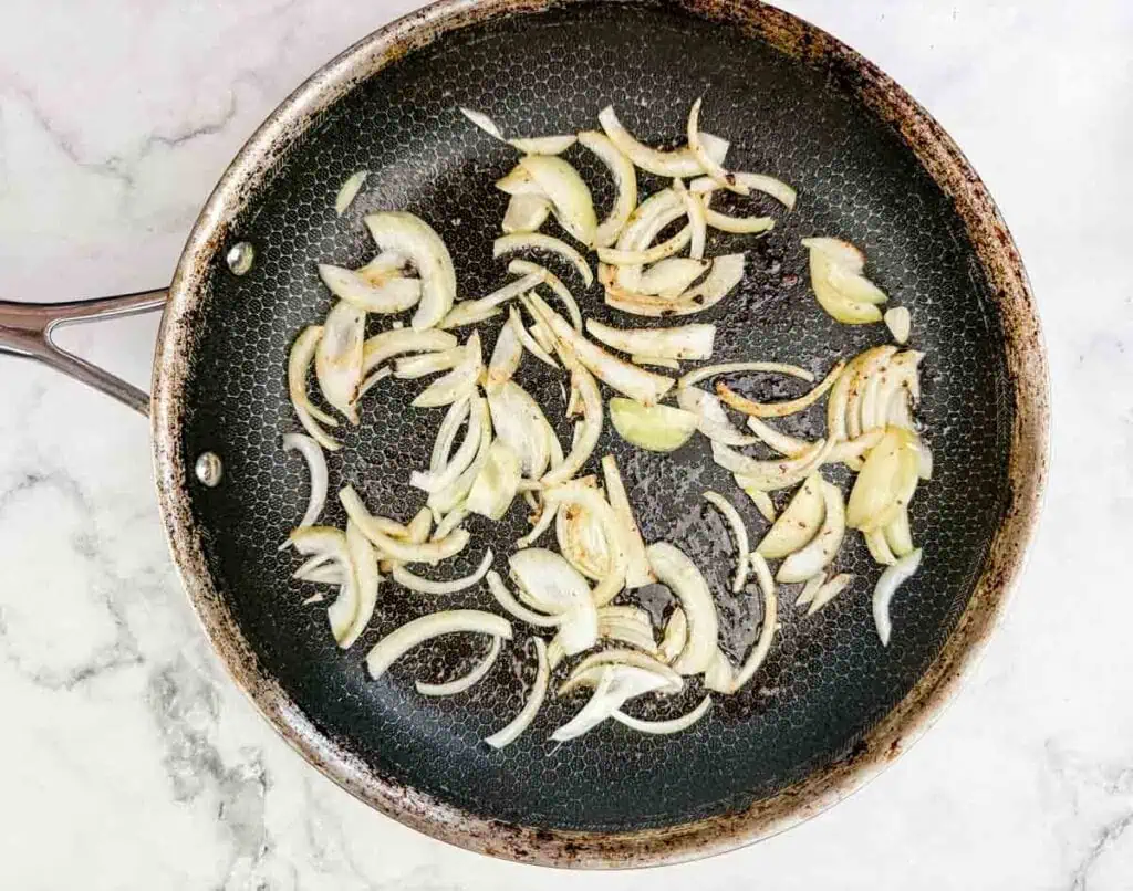 Sliced onions sautéing in a black frying pan on a white marble surface.