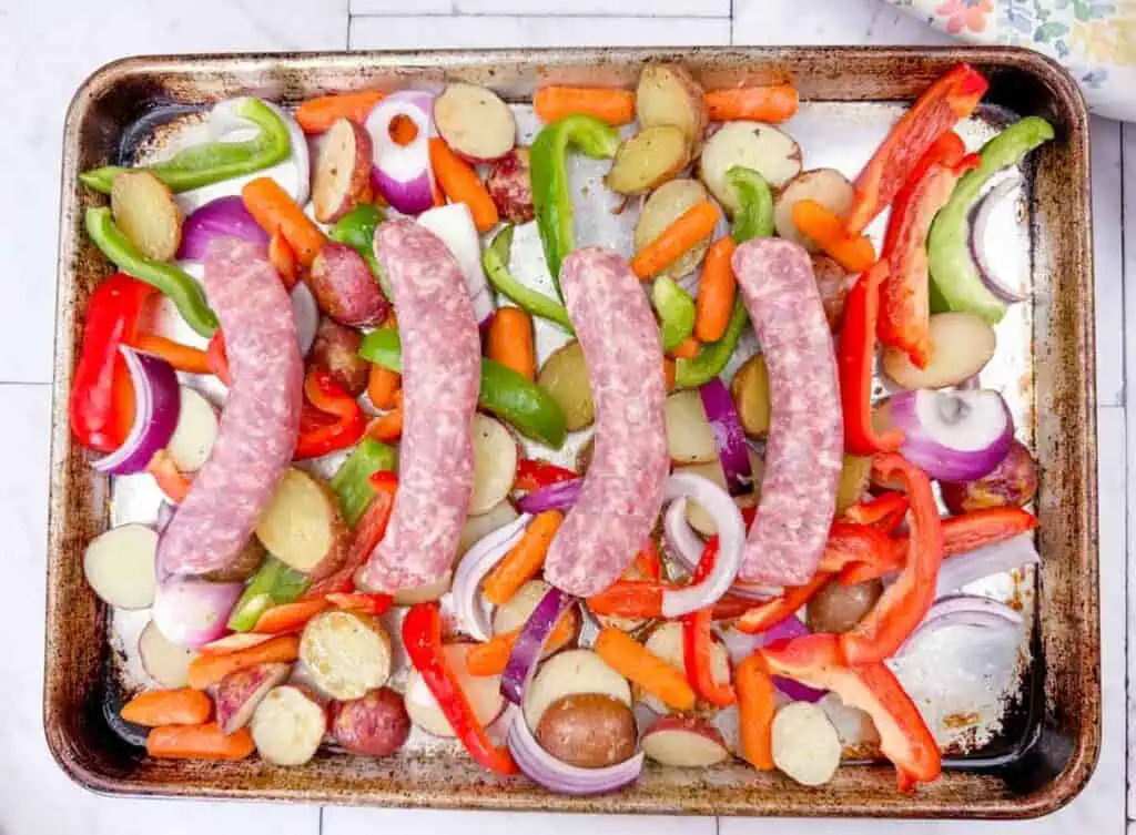 Raw sausages, sliced potatoes, carrots, red onions, and bell peppers arranged on a baking sheet, ready for roasting.