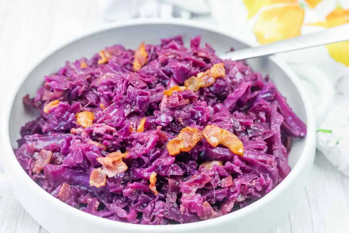 A bowl of cooked red cabbage mixed with small pieces of bacon, served with a spoon.