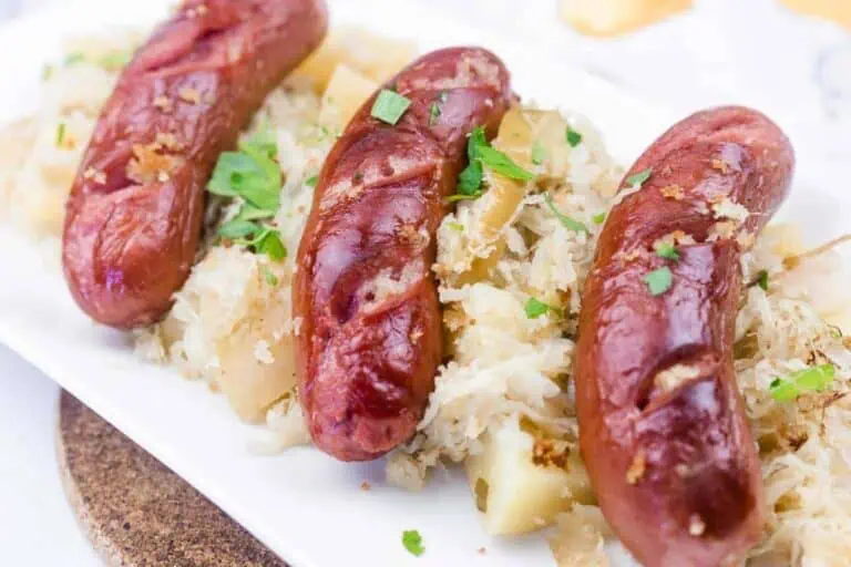 Three grilled sausages served on a bed of sauerkraut and diced potatoes, garnished with chopped parsley, on a white rectangular plate.