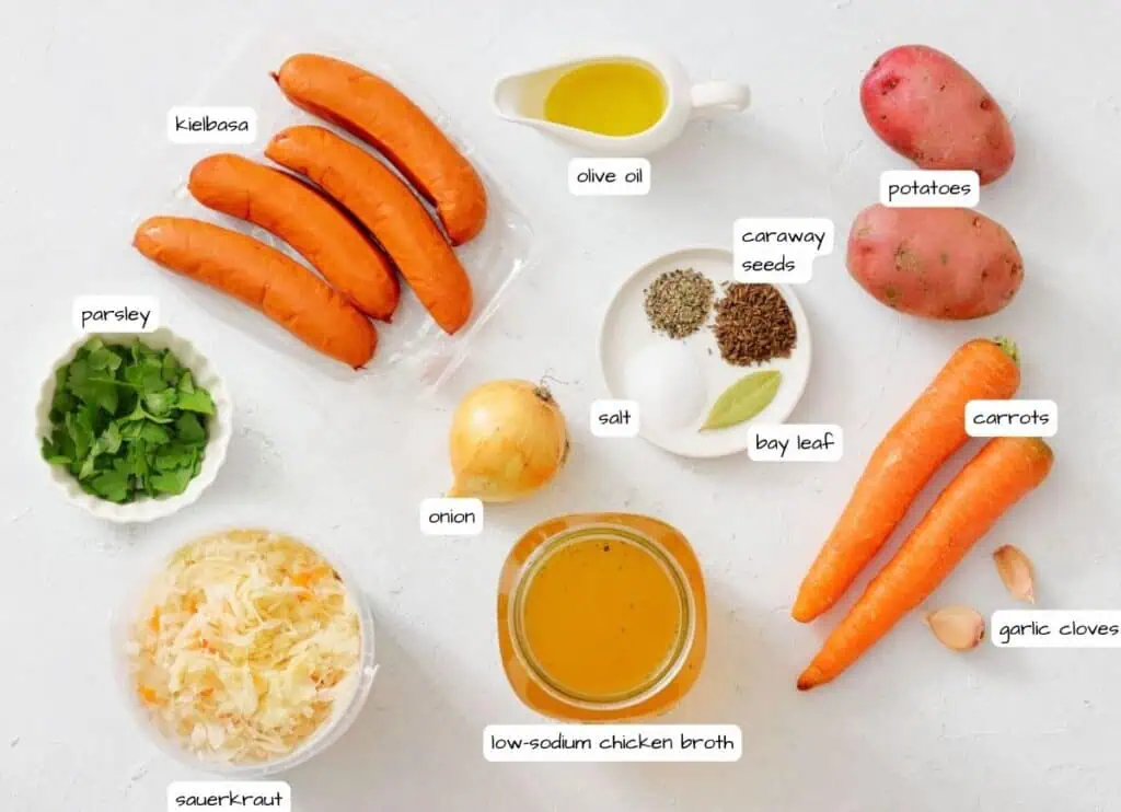 A flat lay of ingredients for a dish, including kielbasa, potatoes, carrots, onion, garlic, sauerkraut, parsley, olive oil, caraway seeds, salt, bay leaf, and chicken broth.