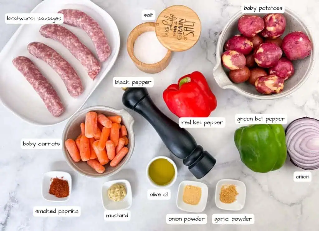 Bratwurst sausages, baby potatoes, baby carrots, red and green bell peppers, red onion, olive oil, mustard, spices, salt, and pepper arranged on a white surface.
