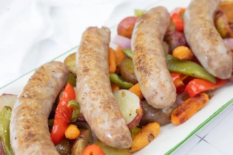Four cooked sausages are served on a platter with roasted potatoes, carrots, red bell peppers, onions, and green peppers.