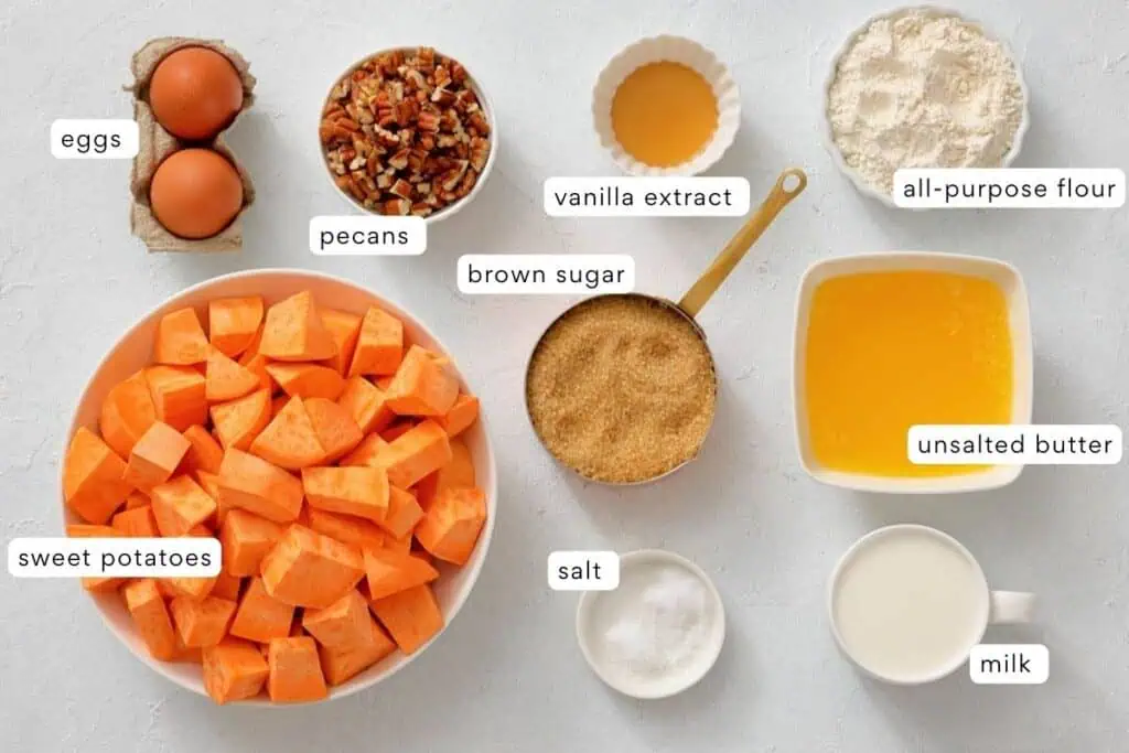 Ingredients for baking: sweet potatoes, eggs, pecans, brown sugar, vanilla extract, all-purpose flour, unsalted butter, salt, and milk arranged on a white surface.