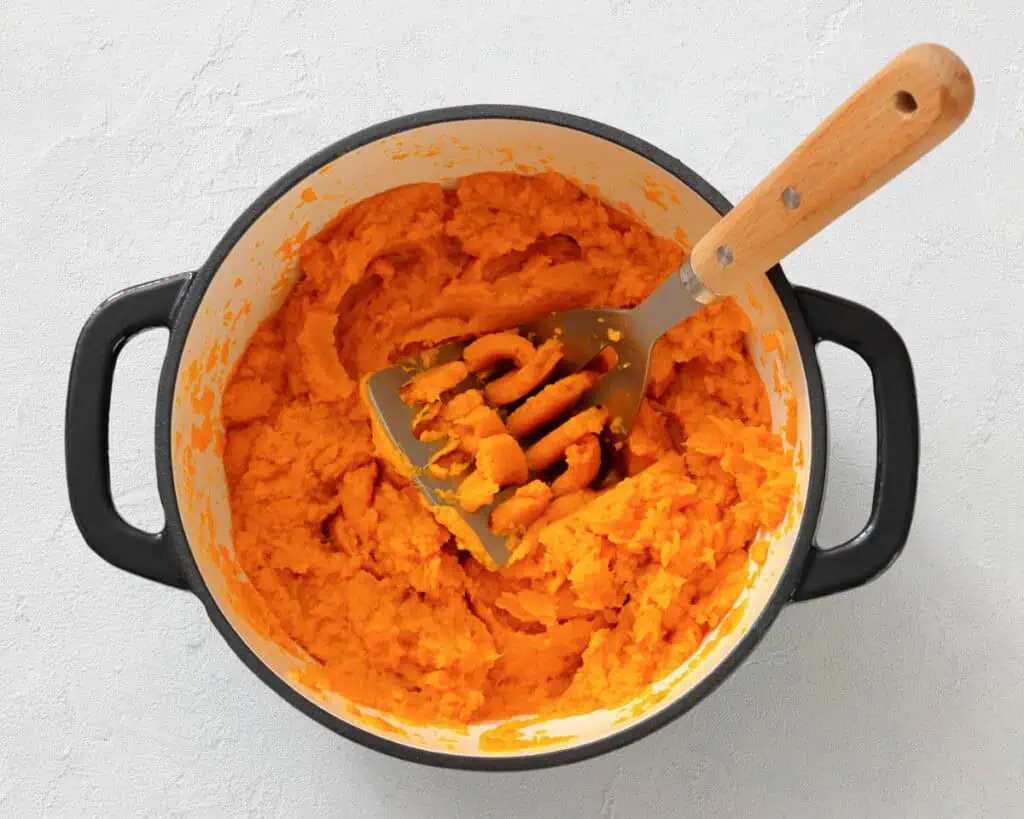 A pot of mashed sweet potatoes with a metal masher resting inside on a light-colored surface.