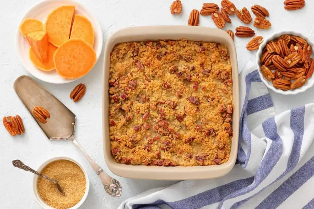 A baked sweet potato casserole topped with pecans in a square dish, surrounded by raw sweet potato slices, pecans, brown sugar, and a striped kitchen towel.