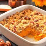 A white baking dish with sweet potato casserole topped with pecan crumble, a corner serving missing, pecans scattered, and measuring cups and a sunflower in the background.