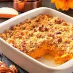 A white baking dish with sweet potato casserole topped with pecan crumble, a corner serving missing, pecans scattered, and measuring cups and a sunflower in the background.