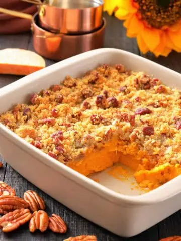 A white baking dish with sweet potato casserole topped with pecan crumble, a corner serving missing, pecans scattered, and measuring cups and a sunflower in the background.