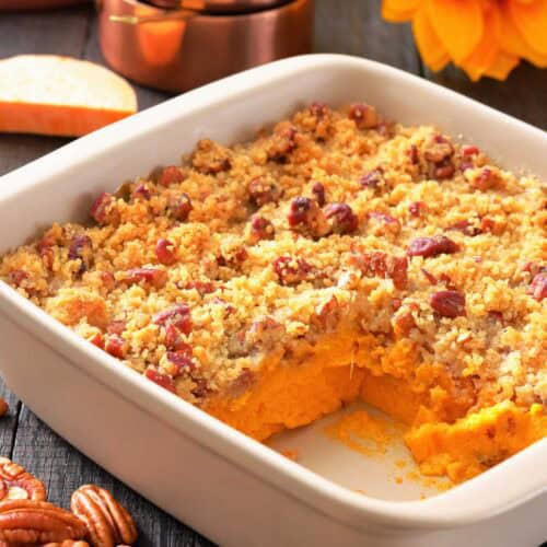 A white baking dish with sweet potato casserole topped with pecan crumble, a corner serving missing, pecans scattered, and measuring cups and a sunflower in the background.