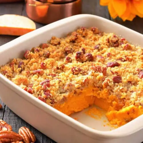 A white baking dish with sweet potato casserole topped with pecan crumble, a corner serving missing, pecans scattered, and measuring cups and a sunflower in the background.