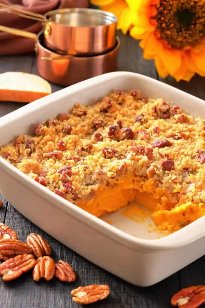 A white baking dish with sweet potato casserole topped with pecan crumble, a corner serving missing, pecans scattered, and measuring cups and a sunflower in the background.