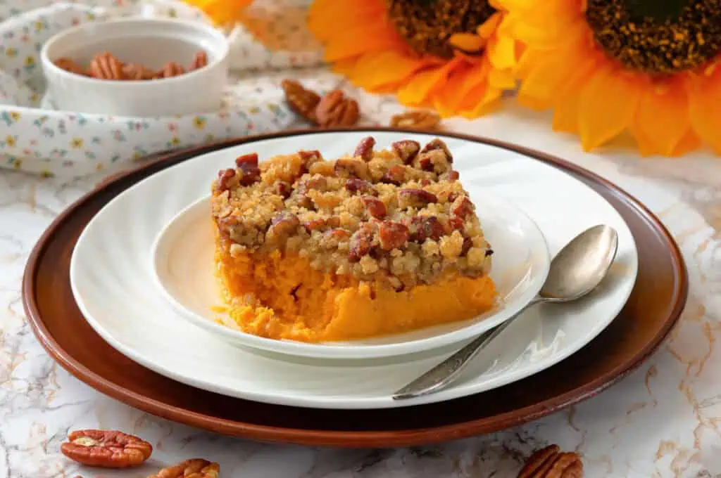 A slice of sweet potato casserole topped with pecan crumble sits on a white plate with a spoon, surrounded by pecans and sunflowers in the background.