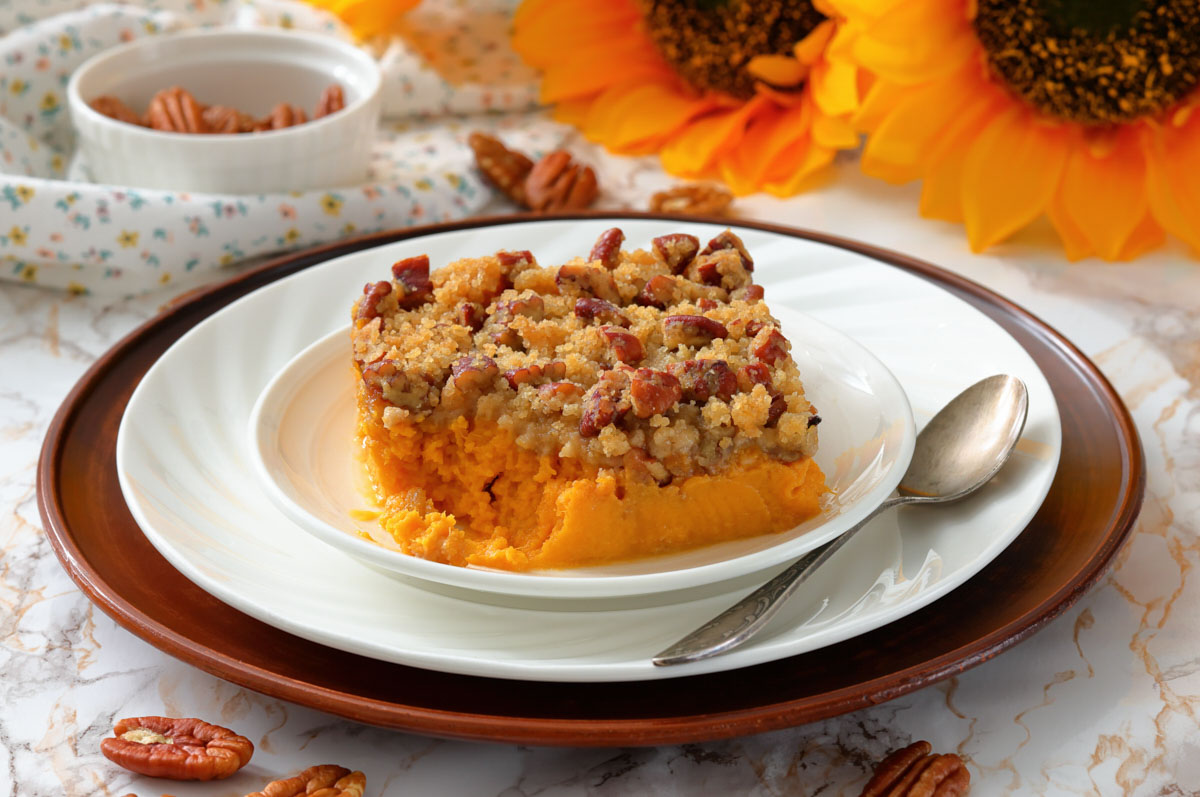 A slice of sweet potato casserole topped with pecan crumble on a white plate with a spoon, surrounded by pecans and sunflowers.