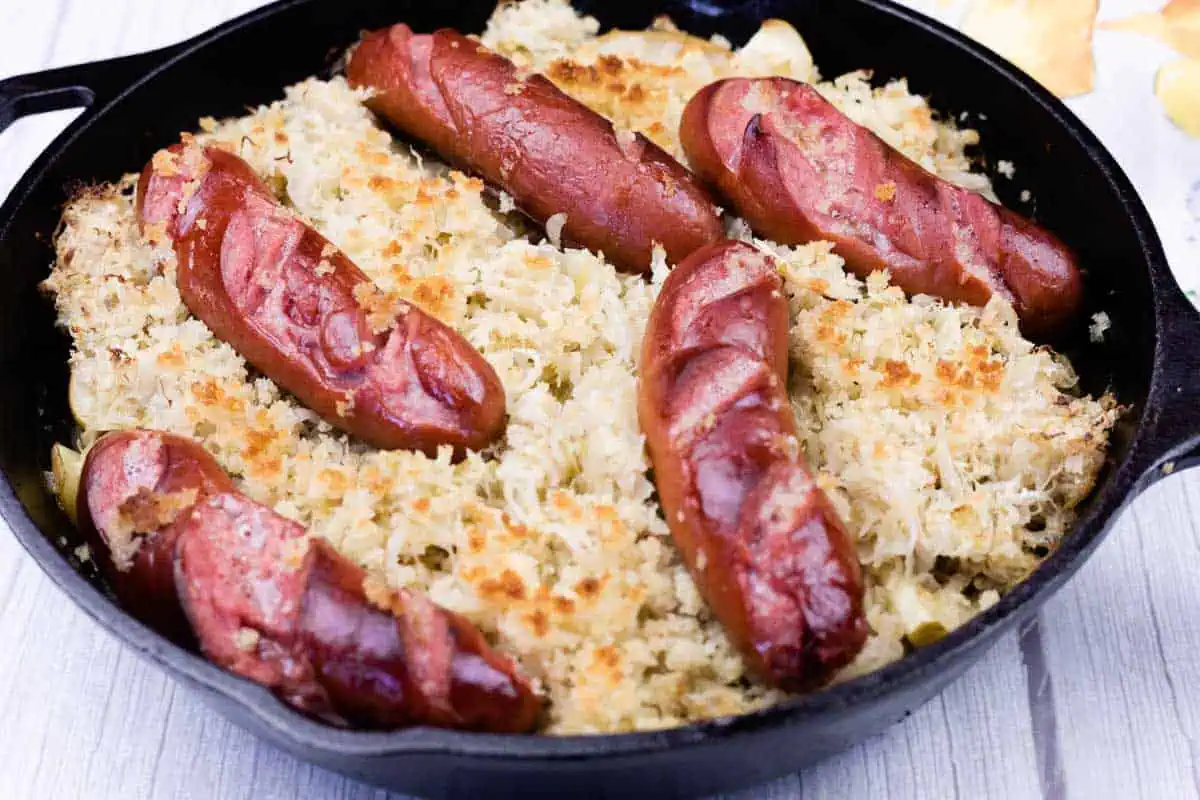 A cast iron skillet filled with sauerkraut and topped with six grilled sausages, all placed on a light wooden surface.
