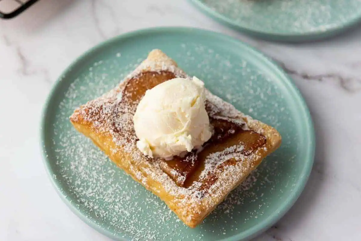A Puff Pastry Apple Tart topped with a scoop of vanilla ice cream and sprinkled with powdered sugar sits on a light green plate.