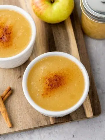 Two bowls of applesauce topped with cinnamon sit on a wooden board, next to an apple, a jar of applesauce, and two cinnamon sticks.