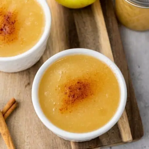 Two bowls of applesauce topped with cinnamon sit on a wooden board, next to an apple, a jar of applesauce, and two cinnamon sticks.