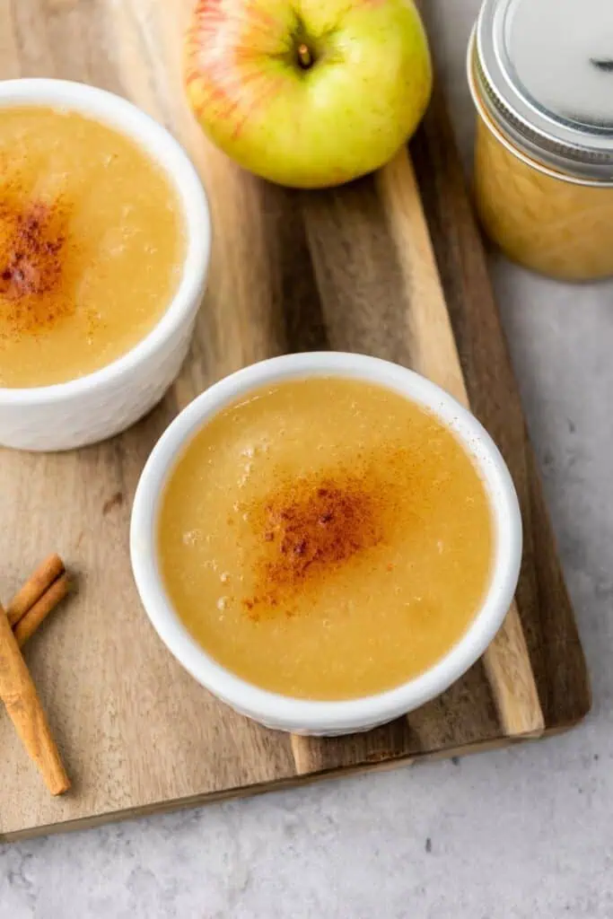 Two bowls of applesauce topped with cinnamon sit on a wooden board, next to an apple, a jar of applesauce, and two cinnamon sticks.