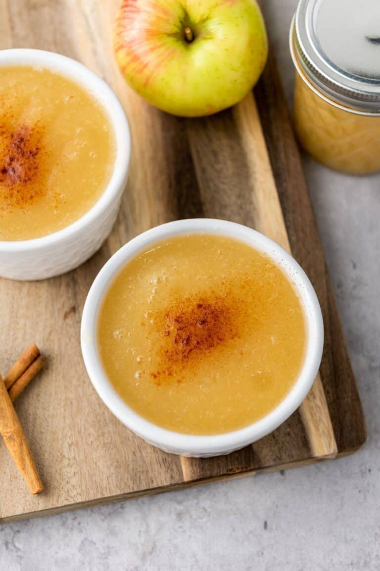 Two bowls of applesauce topped with cinnamon sit on a wooden board, next to an apple, a jar of applesauce, and two cinnamon sticks.