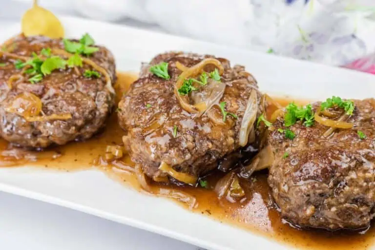 Three hamburger steaks topped with sautéed onions and brown gravy, garnished with chopped parsley, served on a white rectangular plate.