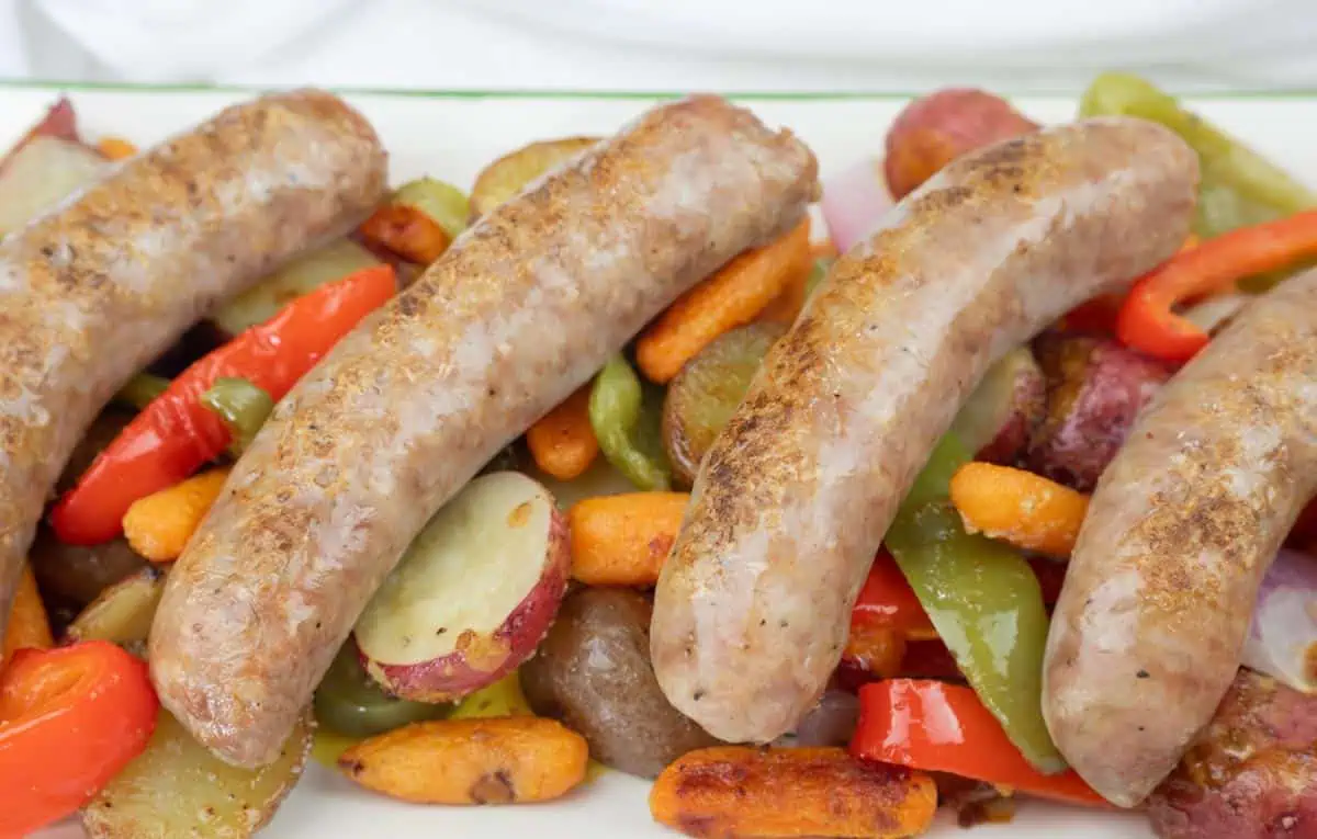 Four cooked sausages served on a bed of roasted vegetables, including carrots, red potatoes, green bell peppers, and red bell peppers.