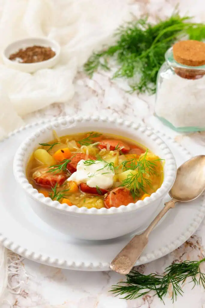 A bowl of vegetable and sausage soup topped with sour cream and fresh dill, accompanied by a spoon and surrounded by salt, pepper, and herbs on a white table.