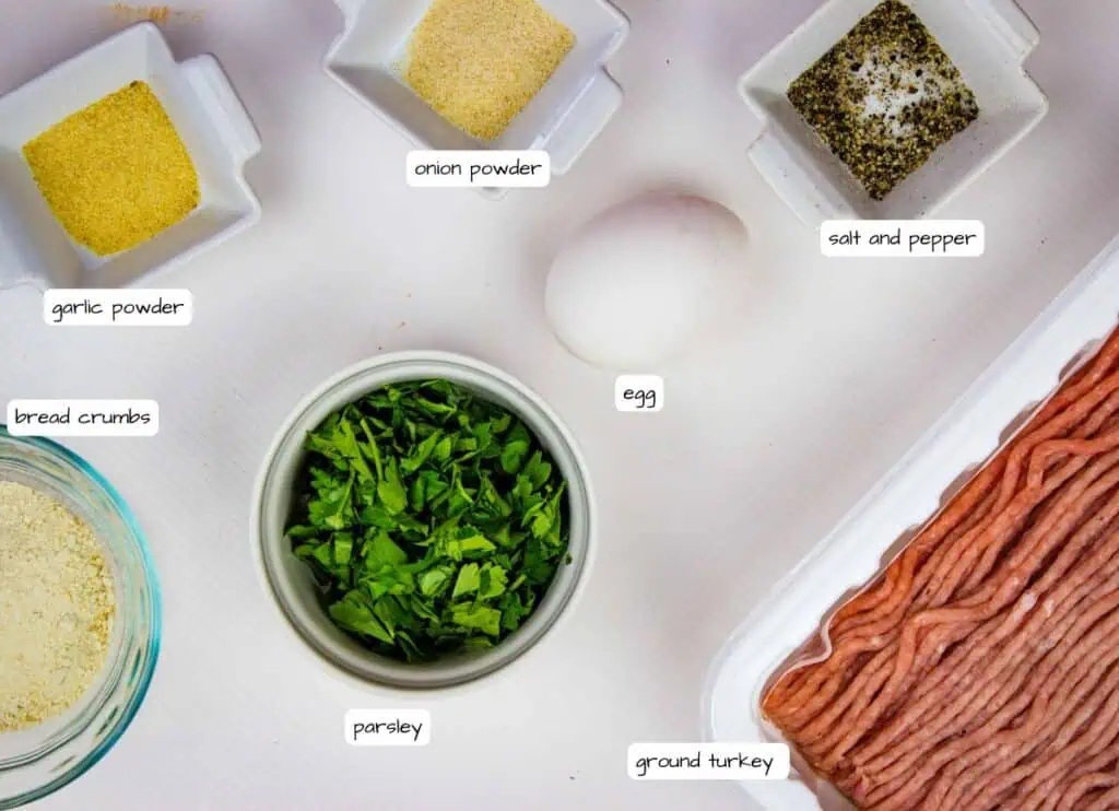 Ingredients for a recipe including ground turkey, an egg, parsley, bread crumbs, garlic powder, onion powder, and salt and pepper displayed in separate containers on a white surface.