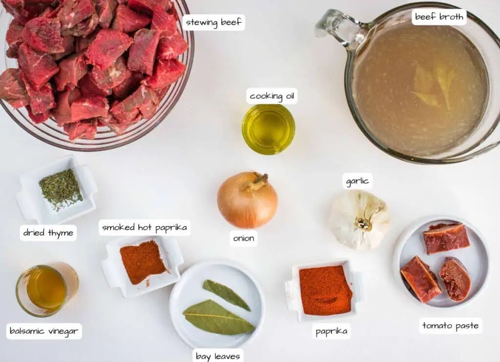 Top-down view of ingredients for a beef stew, including stewing beef, beef broth, onion, garlic, cooking oil, dried thyme, paprika, smoked paprika, bay leaves, tomato paste, and balsamic vinegar.