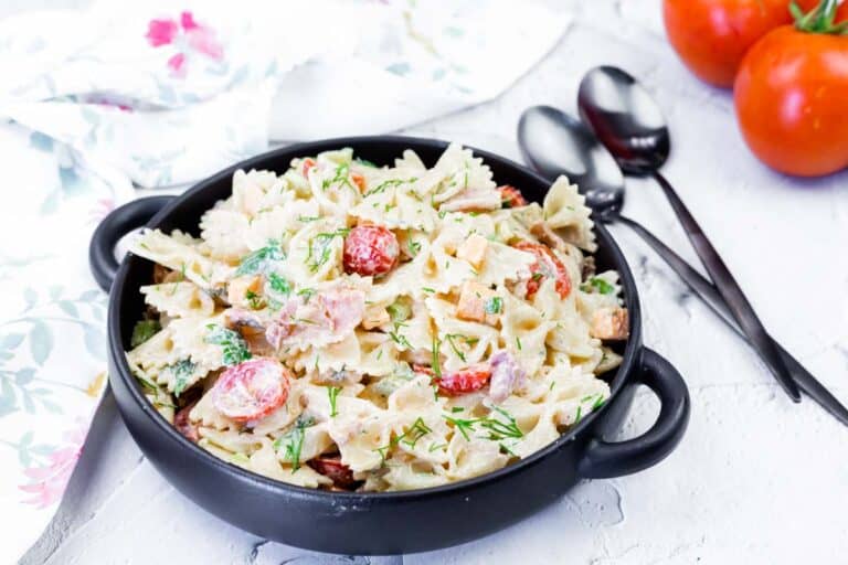 A black bowl filled with creamy bowtie pasta salad mixed with cherry tomatoes, herbs, and vegetables sits on a white surface with two tomatoes and spoons nearby.