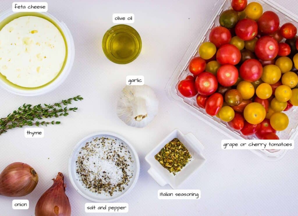 Assorted grape tomatoes, feta cheese, olive oil, garlic, thyme, onion, salt, pepper, and Italian seasoning arranged on a white surface.