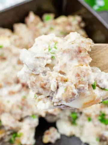A wooden spoon holds a serving of biscuits and gravy with sausage, over a baking dish filled with the same dish, garnished with chopped herbs.